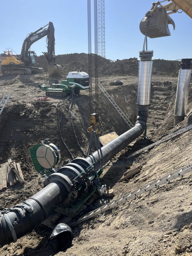 Construction site with large machinery installing metallic pipes underground; excavators and a crane lift and position pipe sections as workers operate equipment in a dirt trench under clear blue sky.
