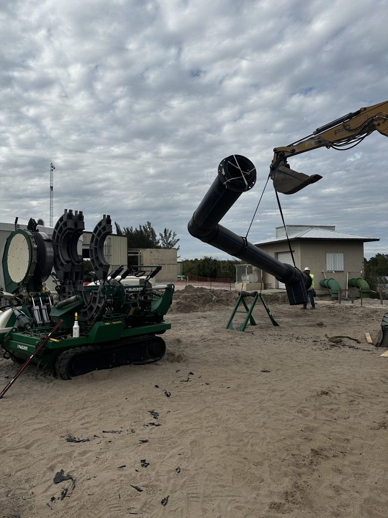 A construction site shows a large excavator lifting a black pipeline section, while workers guide it into position. Nearby, a green fusion machine sits on sandy ground under a cloudy sky. Buildings are visible in the background.