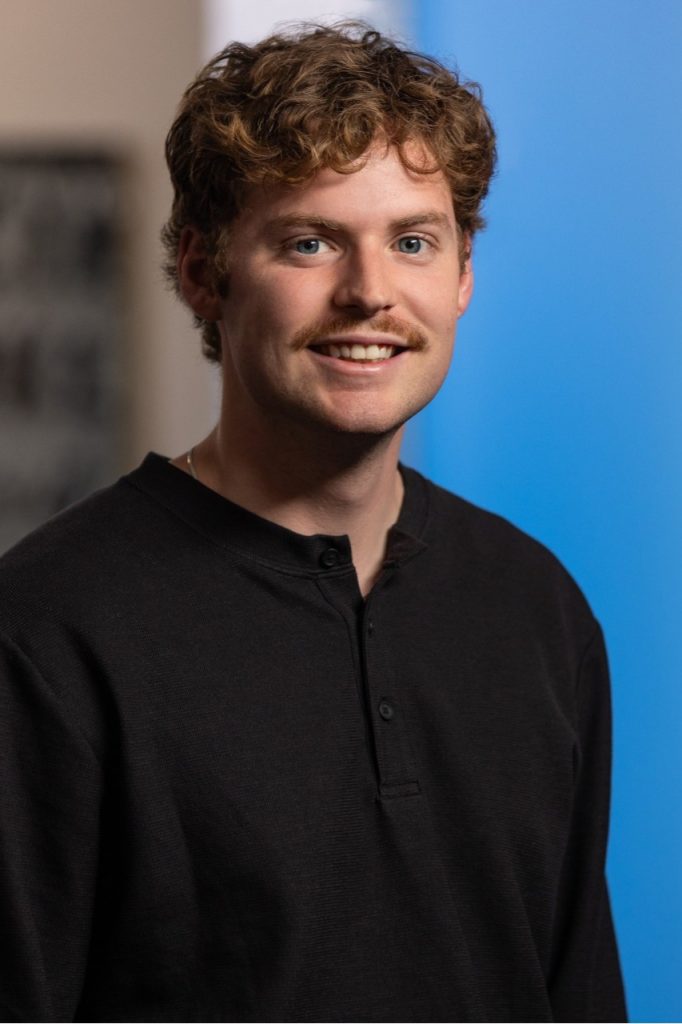 A young man with curly brown hair and a mustache smiles at the camera. He is wearing a black long-sleeve shirt and stands in front of a blurred background with blue and beige tones.