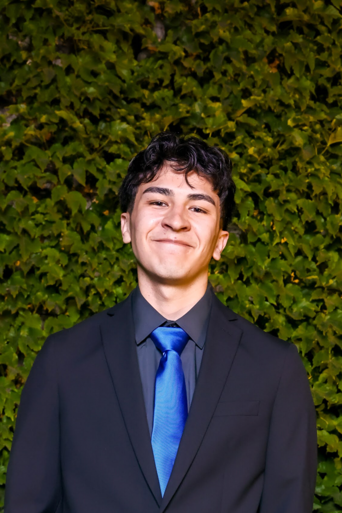 A young man in a black suit, dark shirt, and bright blue tie stands in front of a leafy green background, smiling softly at the camera.