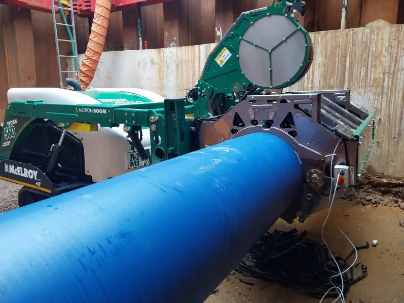 A large blue plastic pipe is being fused using a green McElroy pipe fusion machine at a construction site with concrete walls in the background.