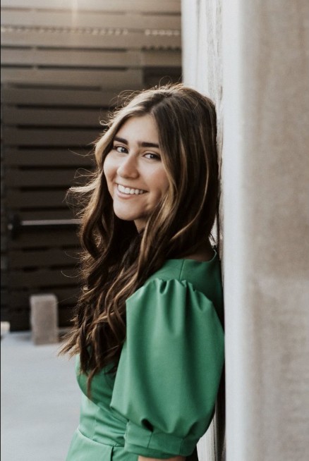 A young woman with long, wavy brown hair wearing a green dress is smiling and leaning against a light-colored wall, with a dark wooden fence in the background.