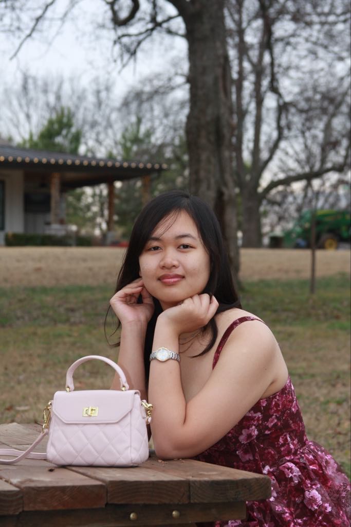 A woman in a floral dress sits at a wooden picnic table outdoors, resting her chin on her hands and smiling. A light pink handbag is on the table. Leafless trees and a house are in the background.