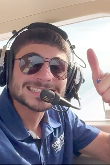 A smiling man wearing sunglasses and a headset gives a thumbs up while sitting in an airplane cockpit. He is wearing a blue shirt with a Spartan logo on it.