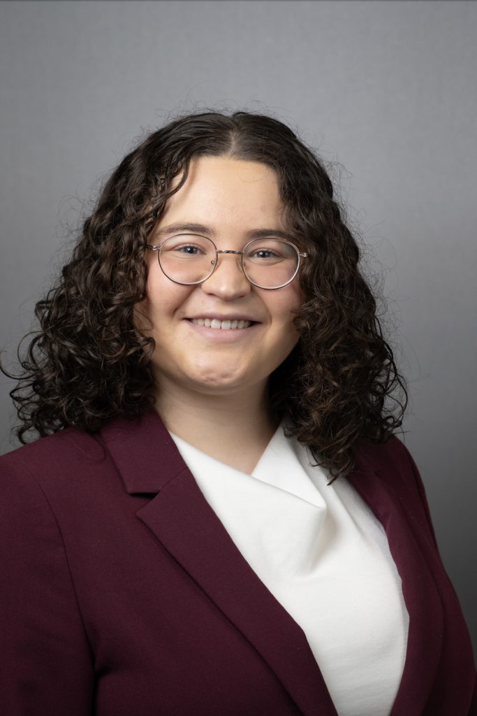 A person with shoulder-length curly brown hair, wearing glasses, a maroon blazer, and a white top, smiles in front of a plain gray background.