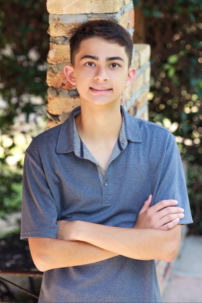 A teenage boy with short dark hair stands outside in front of a brick pillar, smiling with his arms crossed and wearing a gray collared shirt. Green foliage is blurred in the background.