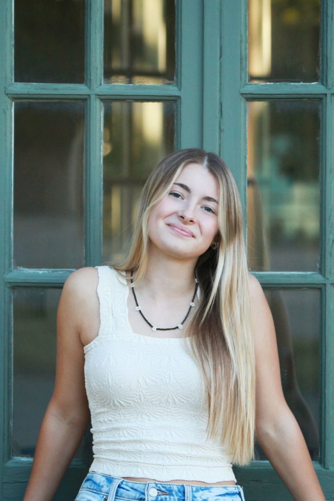 A young woman with long blonde hair, wearing a white sleeveless top and a necklace, stands in front of a green-paned door, smiling gently at the camera.