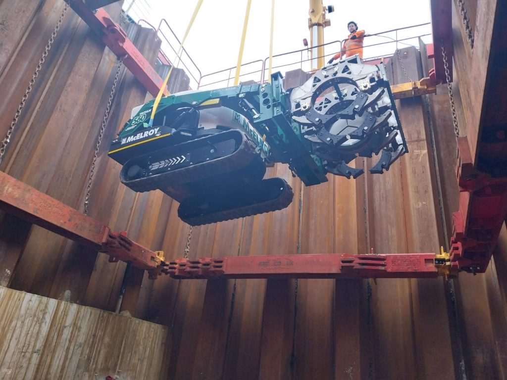 A large drilling machine is being lowered into a deep, metal-lined construction pit by cranes, while a worker in a safety vest observes from above.