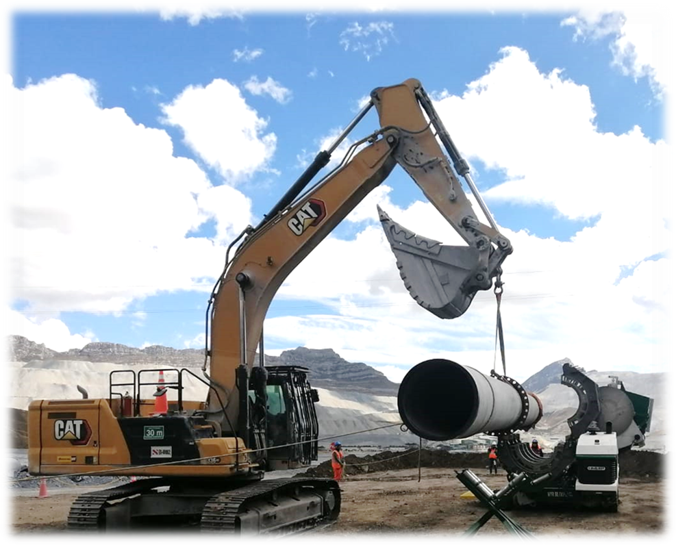 A large yellow excavator lifts a long metal pipe at a construction site with mountains and cloudy sky in the background. Several workers in safety gear are nearby, monitoring the operation.