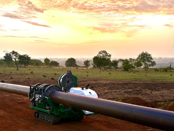 A large machine works on welding or joining a long metal pipeline in a rural landscape at sunrise, with scattered trees and a colorful sky in the background.