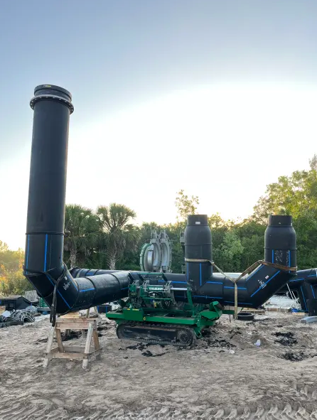 Large black industrial pipes with tall vertical vents are assembled on a sandy construction site, surrounded by trees and a clear sky, with a green machine and scattered materials nearby.