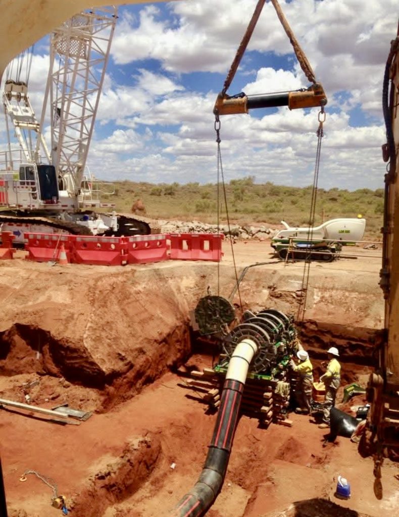 A large crane lowers a heavy pipeline section into a dirt trench as two workers in safety gear guide it. The construction site is set in a dry, open landscape under a partly cloudy sky.