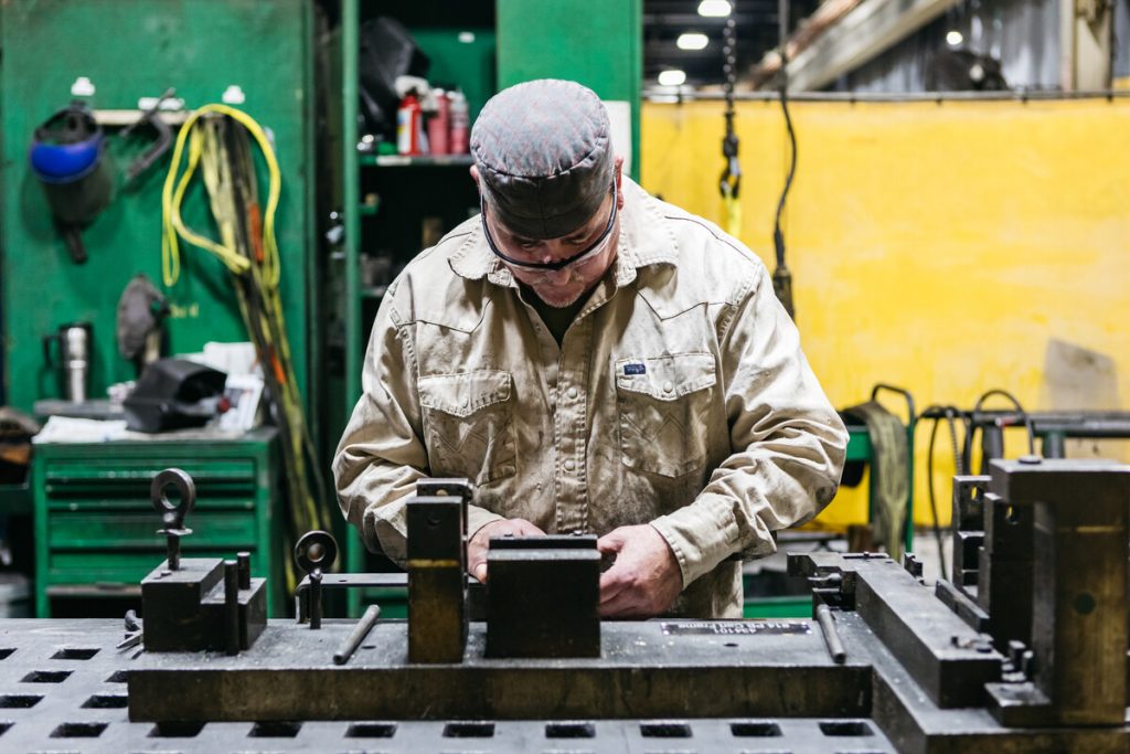 A man wearing safety glasses and a cap works intently at a metal workbench in an industrial workshop, surrounded by tools and machinery.