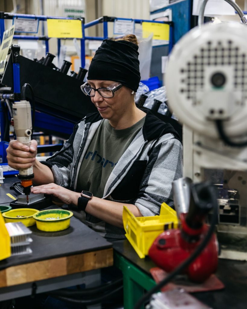 A woman wearing glasses and a beanie uses an electric tool to assemble parts at a workstation in a factory, surrounded by organized bins and machinery.