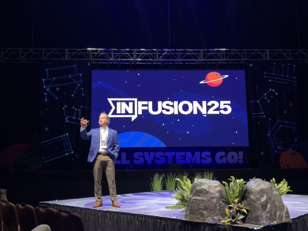A speaker stands on stage in front of a large screen displaying INFUSION25 with a planet graphic. Stage decorations include rocks, plants, and illuminated designs. The words ALL SYSTEMS GO! are visible below the screen.