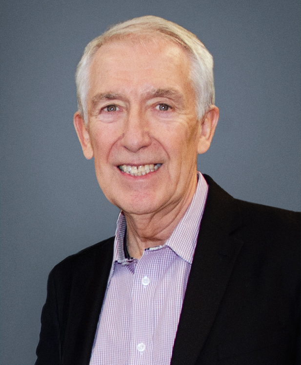 An older man with short gray hair is smiling at the camera. He is wearing a light purple checked shirt and a black blazer, posed against a plain gray background.