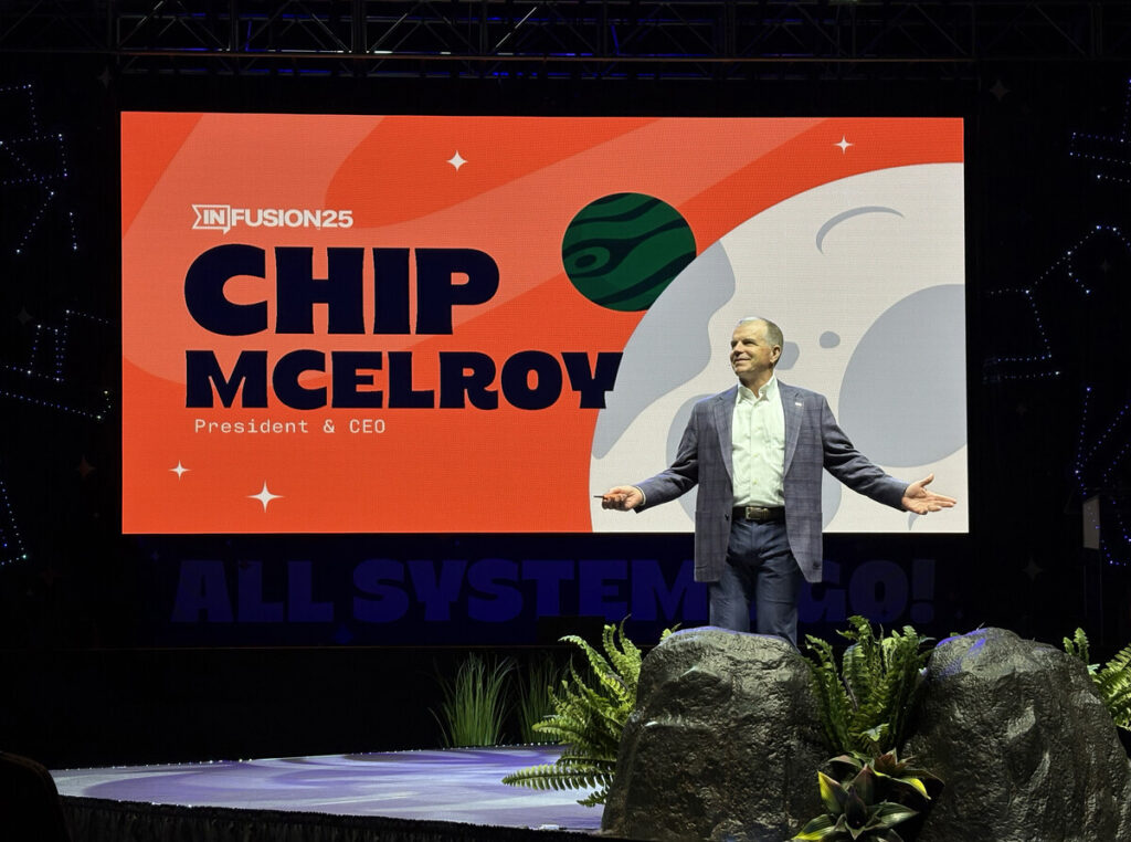 A man in a suit stands on stage in front of a colorful screen that reads INFUSION25, CHIP MCELROY, President & CEO. The stage has rocks and plants in the foreground.