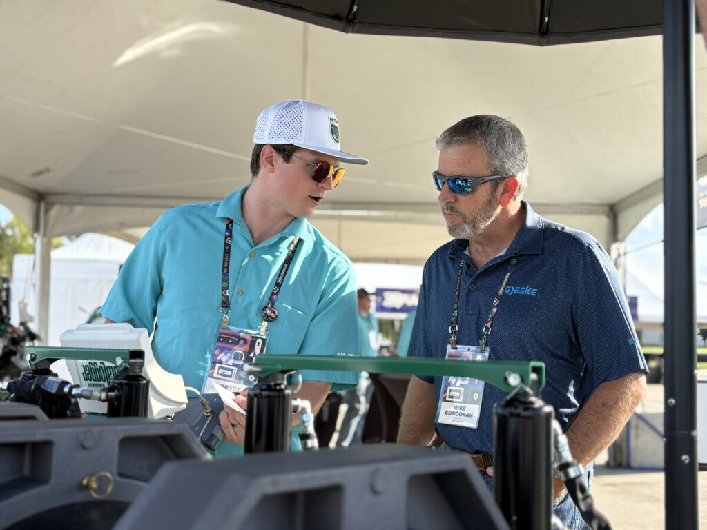 Two men wearing sunglasses and event badges stand under a tent, engaged in conversation while looking at equipment on display at an outdoor event. One man points at the equipment as they discuss.