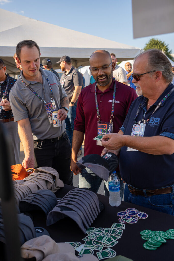 Three men smile and interact at an outdoor event booth, examining hats and trading chips on a table covered with hats and patches. Other people and a white tent are visible in the background.