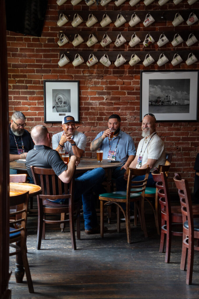 Five men sit around a table in a cozy pub with brick walls, drinking beer and talking. The background features teapots displayed on shelves and black-and-white framed photos on the wall.