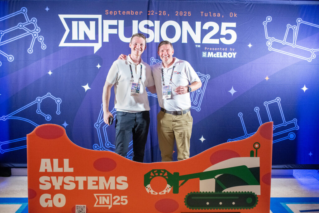 Two smiling men stand together posing for a photo at the IN Fusion 25 event in Tulsa, Oklahoma, with a colorful backdrop and a sign that reads ALL SYSTEMS GO IN25 featuring construction graphics.