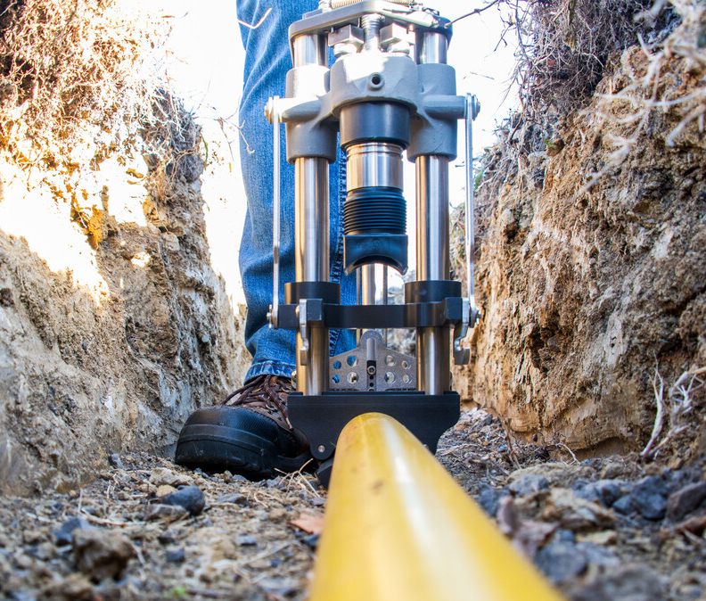 Close-up view of a trench with a yellow pipe being installed and a person operating a large, metal pipe-laying tool, visible from ground level. The person wears jeans and work boots.