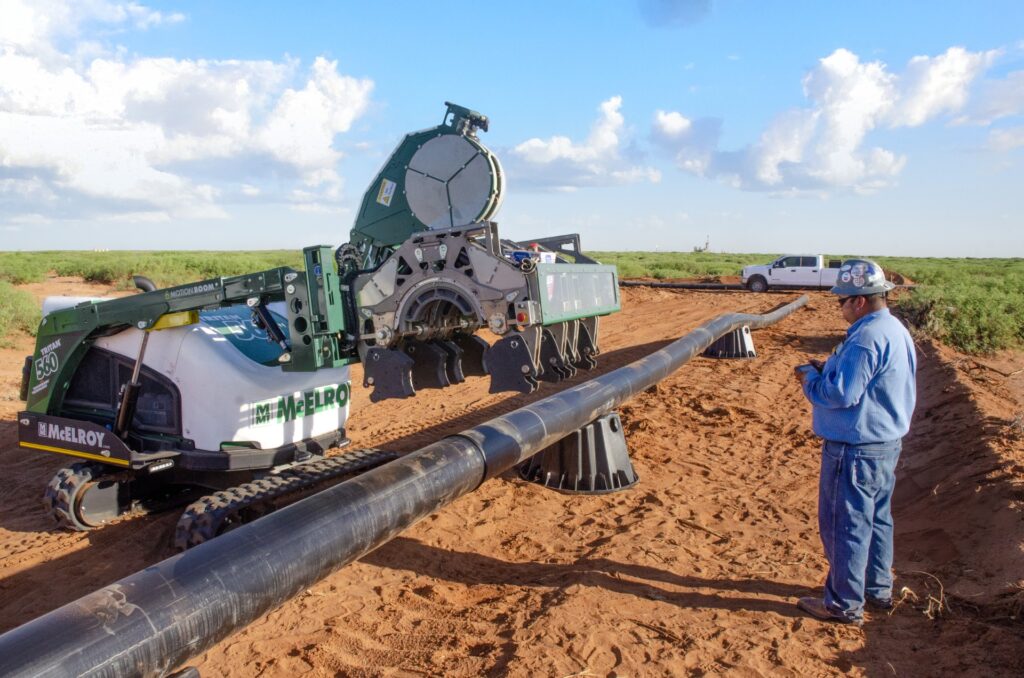 A worker in a blue outfit monitors a large machine as it prepares to fuse long black plastic pipes on a sandy construction site under a blue sky with scattered clouds. A white pickup truck is parked in the background.