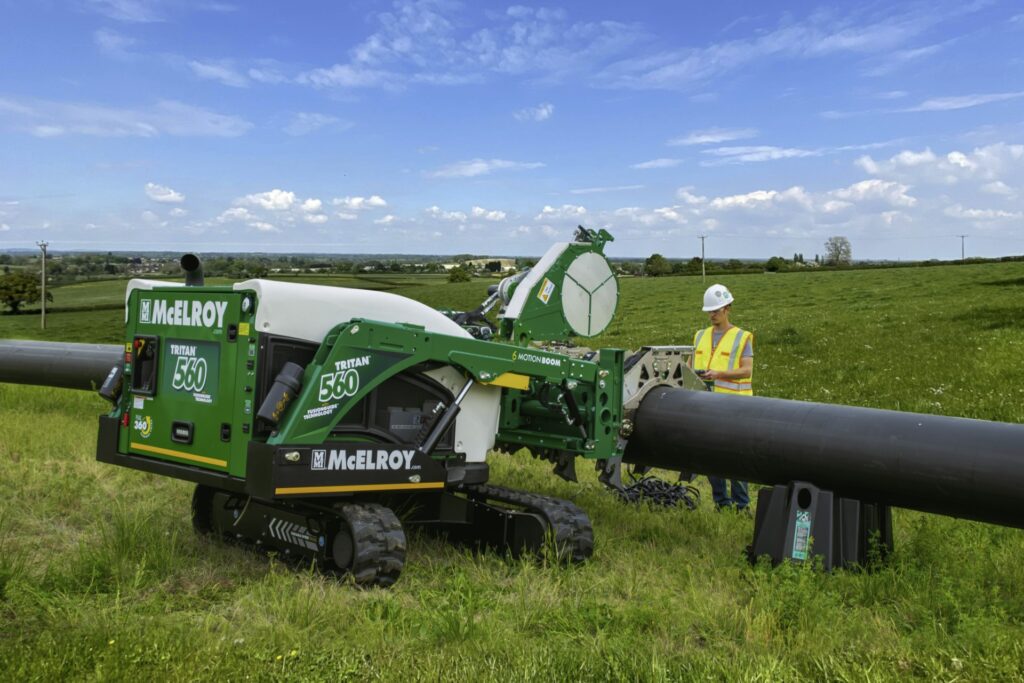 A worker in a safety vest and helmet operates a large green McElroy pipe fusion machine labeled TracStar 630 on grassy terrain, aligning two black pipelines in an open field under a partly cloudy sky.