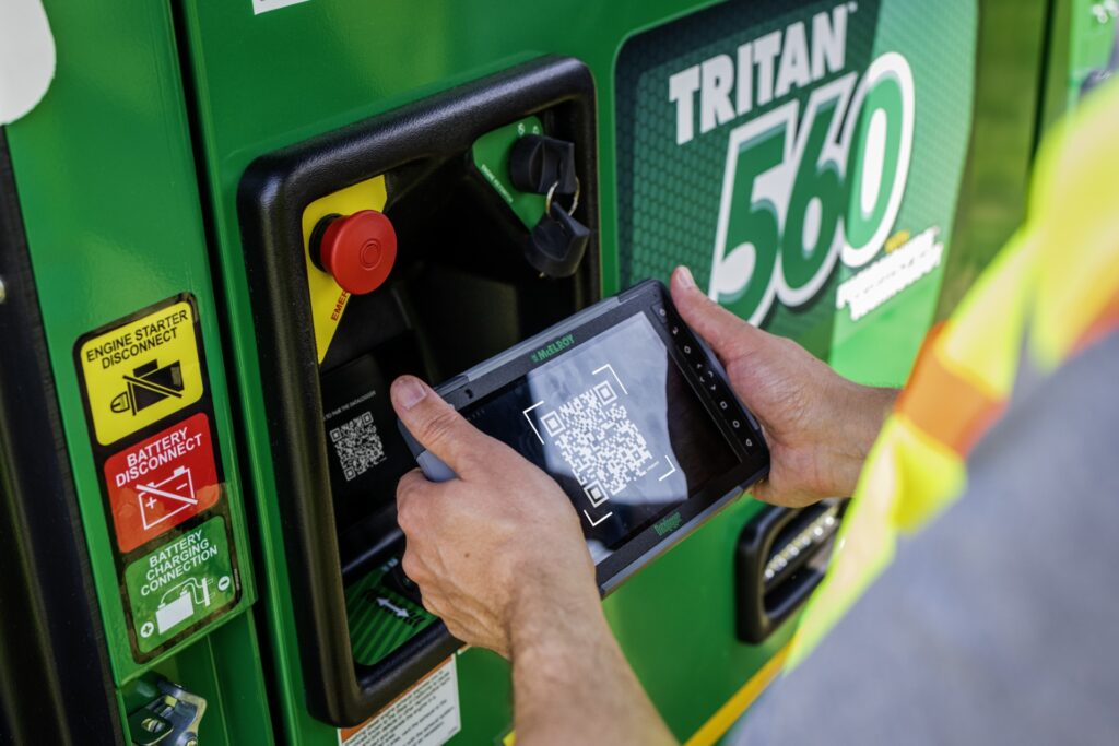 A person scans a QR code on green machinery labeled TRITAN 560 using a handheld device. Visible are safety labels, including Engine Starter Disconnect and Battery Disconnect.