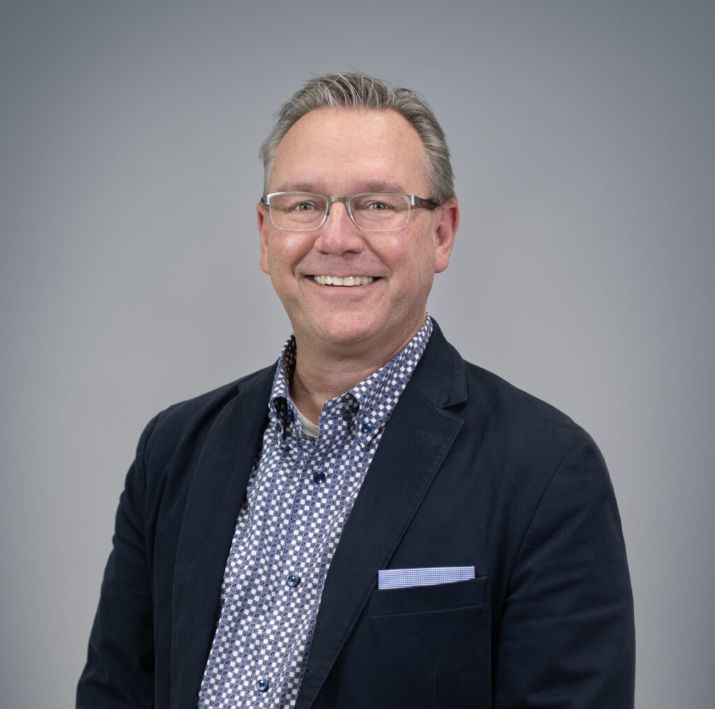 A smiling man with short, gray hair and glasses wearing a navy blazer over a patterned shirt stands in front of a plain gray background.