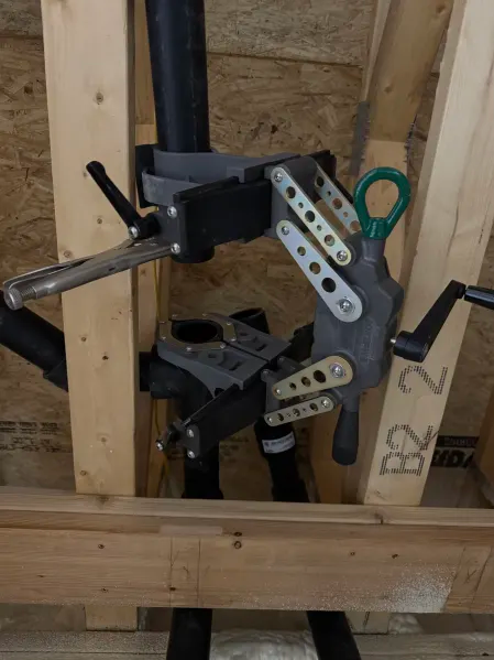 A pipe welding alignment clamp is attached to black pipes within a wooden wall frame, helping hold the pipes in place for precise fitting and welding.