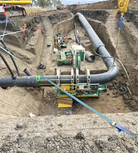 A large pipeline is being welded and assembled in a trench, with heavy machinery and equipment surrounding the pipe. The area is marked off with caution tape and the ground is mostly dirt.