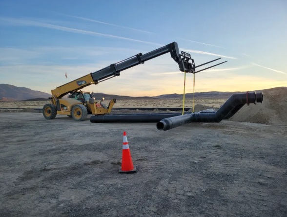 A forklift lifts a large black pipe at a construction site in a desert landscape during sunset. An orange traffic cone is in the foreground, and mountains are visible in the distance.