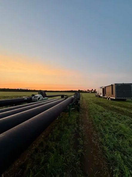 Large black pipes are laid out across a grassy field at sunset, with trucks and trailers parked on the right side under a clear sky transitioning from orange to blue.