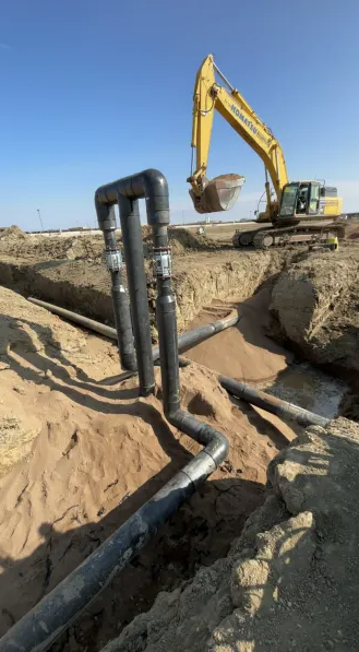 A yellow excavator works at a construction site with exposed black pipes in sandy soil under a clear blue sky. The pipes are arranged vertically and horizontally in the trench.