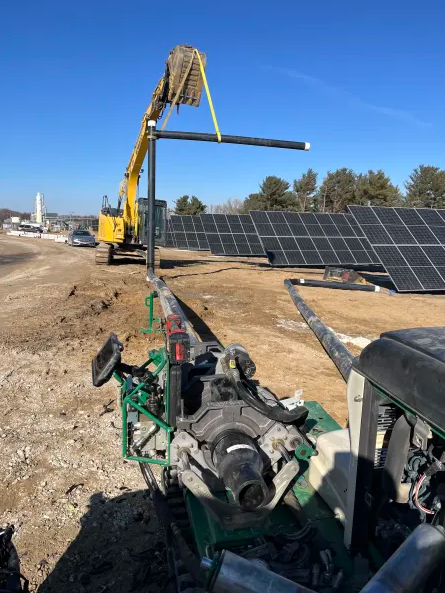 A construction site where a yellow excavator lifts a metal pipe near rows of solar panels, with machinery and tools in the foreground under a clear blue sky.