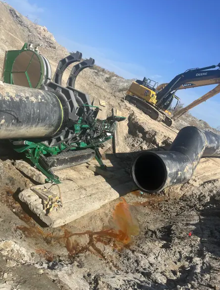 A large construction site with two big black pipes, one on the ground and one held by green machinery. An excavator is parked in the background on sandy, uneven terrain under a clear blue sky.