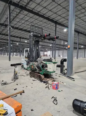 A warehouse forklift is parked indoors with pipes and equipment parts scattered around it. The forklift appears to be undergoing repairs or modifications. The large, open warehouse is mostly empty.