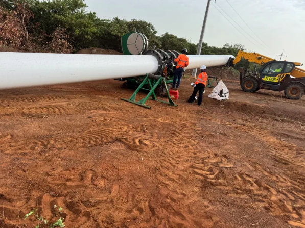 Two workers in safety gear inspect and work on a large wind turbine blade lying on the ground at a construction site, with machinery and green vegetation in the background.