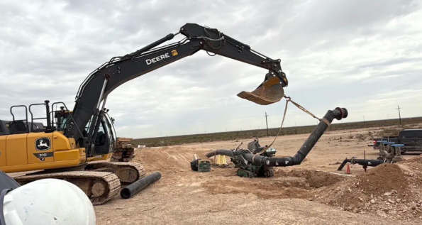 A John Deere excavator lifts a large black pipe into place at a construction site in a dry, open landscape under a cloudy sky. Other pipes and machinery are visible nearby.