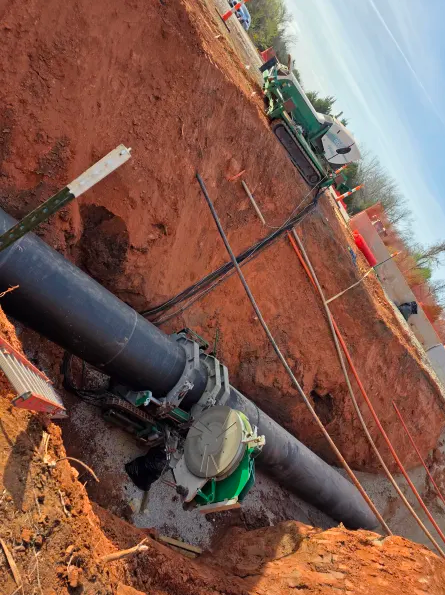A large black pipe is being fused underground at a construction site. Heavy equipment surrounds the pipe, and orange safety barriers are set up around the excavation area.