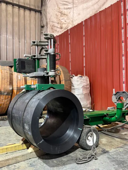 A large black metal pipe is held in place by a green industrial cutting or drilling machine inside a workshop with red metal walls and various equipment in the background.