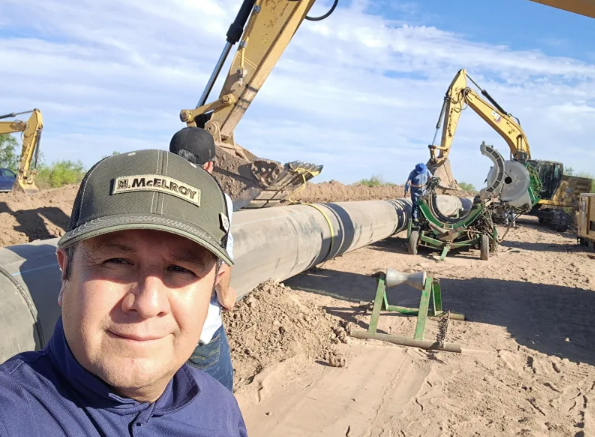 A man in a cap takes a selfie at a construction site where workers and excavators are installing a large pipeline in a sandy area under a blue sky.