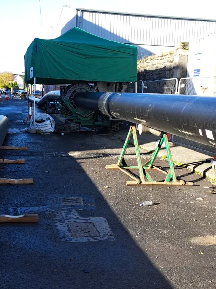 A large black pipeline is supported by metal stands on a paved surface, leading to a green canopy tent with construction equipment underneath. Construction materials and barriers are visible in the background.