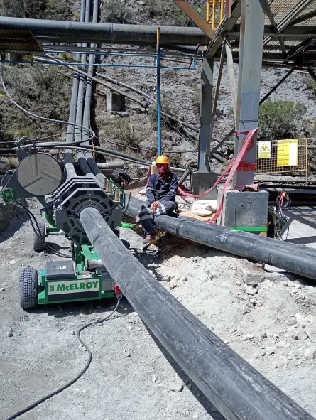 A worker wearing safety gear operates a McElroy pipe fusion machine, joining large black pipes at an industrial outdoor site with rocky ground and metal structures in the background.