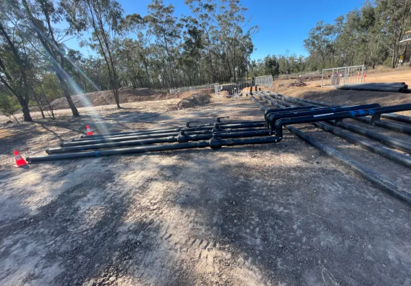 Several large black pipes are laid out on bare soil at a construction site, surrounded by trees and some fencing. Traffic cones are placed near the pipes under a clear blue sky.