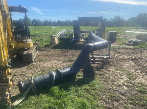 A large black pipeline with multiple bends lies on the ground at a construction site. A yellow excavator is parked nearby, and various equipment and supplies are visible in the background on a grassy field.
