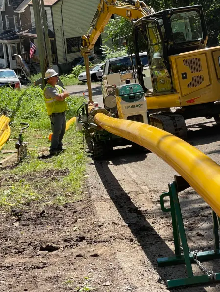 A construction worker in a safety vest and helmet stands next to a large yellow pipe being moved by an excavator on a residential street, with houses and greenery in the background.