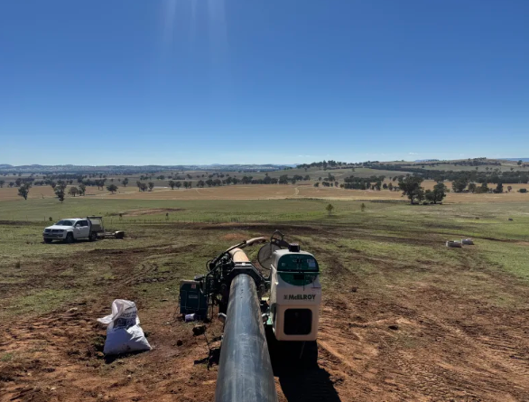 Large pipeline construction equipment and a long pipe stretch across an open grassy field, with a white truck parked nearby. The landscape is expansive, under a clear blue sky.