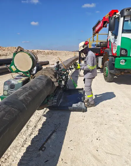A worker in safety gear operates machinery to weld large black pipes outdoors on a sandy construction site, with a green and white service truck parked nearby under a blue sky.